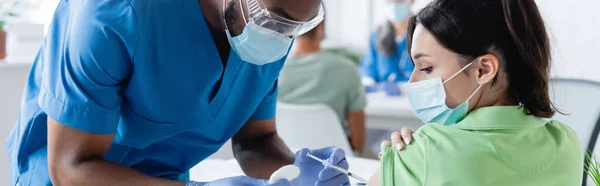 African american doctor in medical mask vaccinating young woman in hospital, banner — Stock Photo