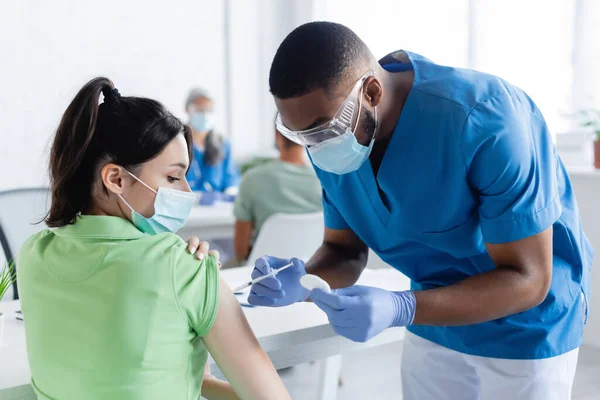 Médecin afro-américain sous masque médical vaccinant une jeune femme en clinique — Photo de stock