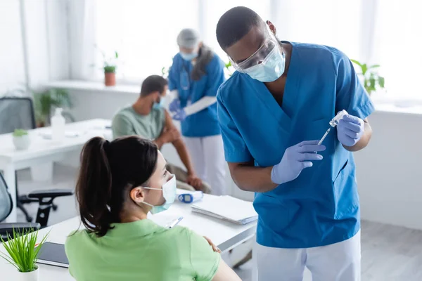 Médico afro-americano preparando vacina perto de mulher em máscara médica e colega turvo com paciente — Stock Photo