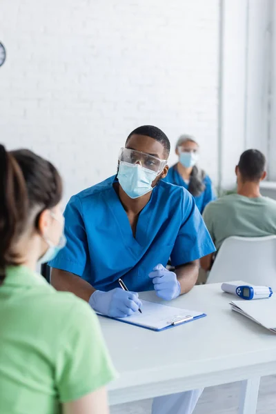 Medico afroamericano con appunti e penna che parla con una donna in maschera medica, concetto di vaccinazione — Foto stock