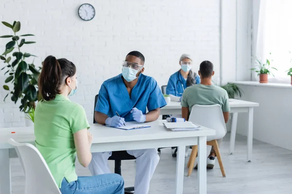 Medici interrazziali in maschere mediche che parlano con i pazienti nel centro di vaccinazione — Foto stock