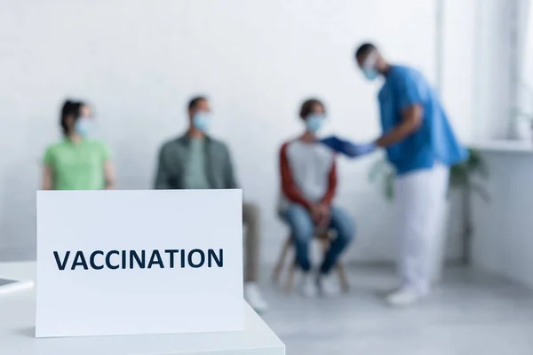 Placa de sinal com letras de vacinação perto de médico afro-americano desfocado e pacientes — Fotografia de Stock
