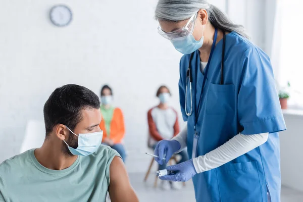 Mature asiatique médecin dans masque médical tenant seringue et pot avec vaccin près de jeune homme et les patients flous — Photo de stock