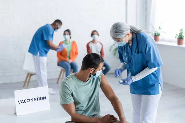 Mature asiatique infirmière tenue vaccin près des hommes dans masque médical tandis que flou afro-américain collègue parler avec patients — Photo de stock