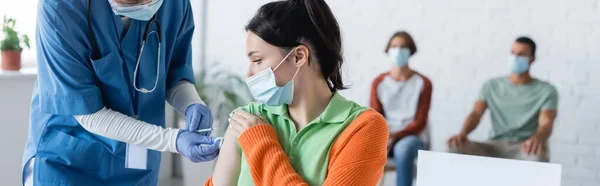 Infirmière en gants de latex vaccinant la jeune femme en masque médical près des patients flous, bannière — Photo de stock