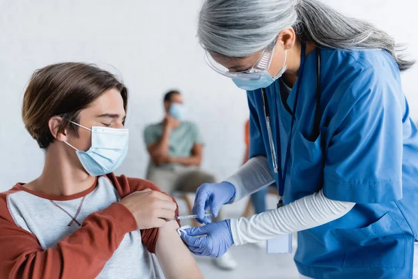 Maduro asiático en médico máscara vacunación joven paciente cerca borrosa africano americano hombre - foto de stock