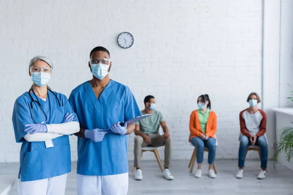 Médicos inter-raciais em máscaras médicas e óculos olhando para a câmera perto de pacientes em fundo borrado — Fotografia de Stock