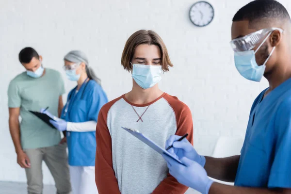 Полиэтнические врачи в медицинских масках пишут на планшетах рядом с пациентами в центре вакцинации — стоковое фото