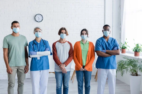 Médicos interraciales y pacientes con máscaras médicas mirando a la cámara en el centro de vacunación - foto de stock