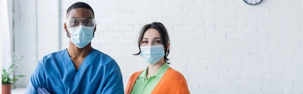 Mujer joven y médico afroamericano en máscaras médicas mirando a la cámara, concepto de vacunación, pancarta - foto de stock