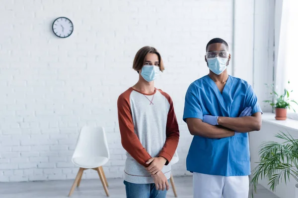 Африканский американский врач, стоящий со скрещенными руками рядом с молодым человеком в медицинской маске, концепция вакцинации — стоковое фото