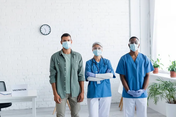 Médicos multiétnicos y hombre joven con máscaras médicas mirando a la cámara en el centro de vacunación - foto de stock