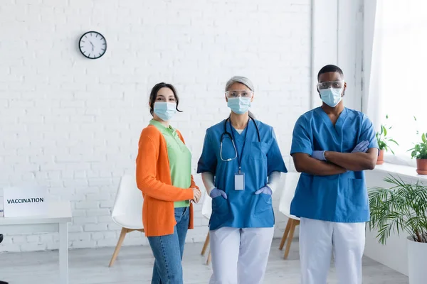 Junge Frau und multiethnische Ärzte mit medizinischen Masken und Schutzbrillen blicken in die Kamera im Impfzentrum — Stockfoto