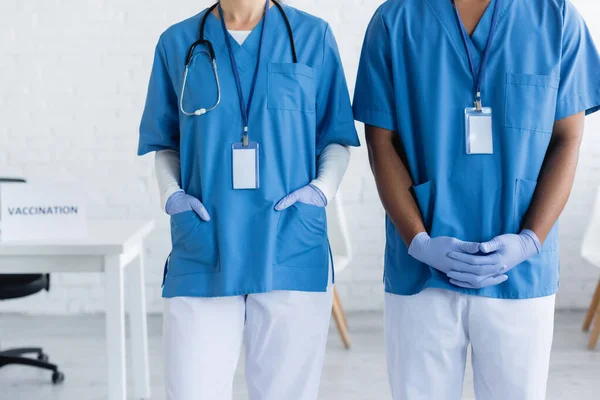 Vista recortada de los médicos interracial en uniforme con etiquetas de nombre en el centro de vacunación - foto de stock