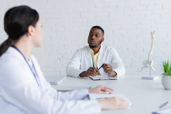 Afrikanischer amerikanischer Arzt hält Stift in der Hand, während er mit Kollegen über verschwommenen Vordergrund spricht — Stockfoto