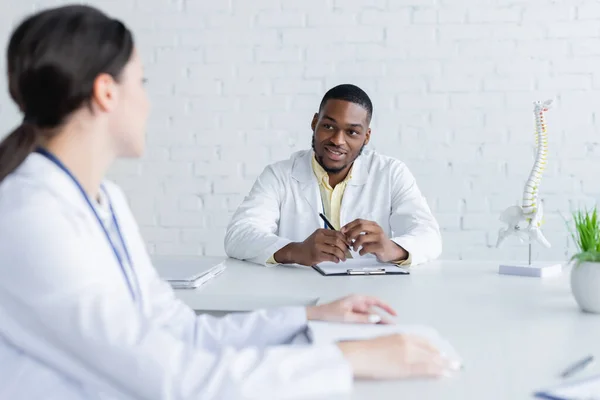 Sonriente afroamericano médico sentado cerca de columna vertebral modelo y hablando con borrosa colega - foto de stock