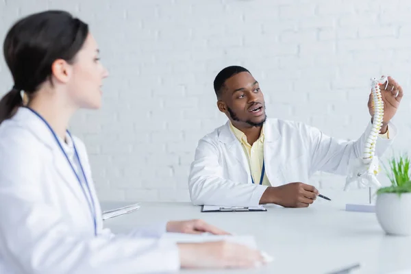 African american doctor pointing at spine model while talking to blurred colleague — Stock Photo