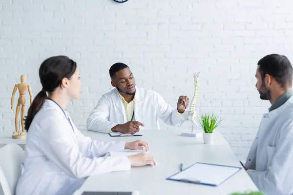 African american doctor pointing with pen at spine model during council with colleagues — Stock Photo