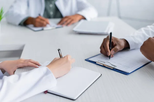 Cropped view of multiethnic doctors writing during council in meeting room — Stock Photo