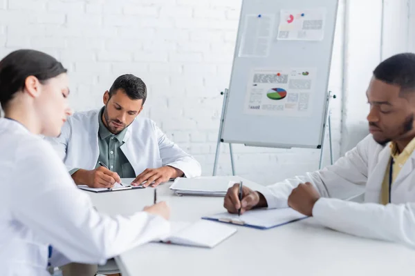 Multicultural doctors writing in conference room near flip chart with infographics — Stock Photo