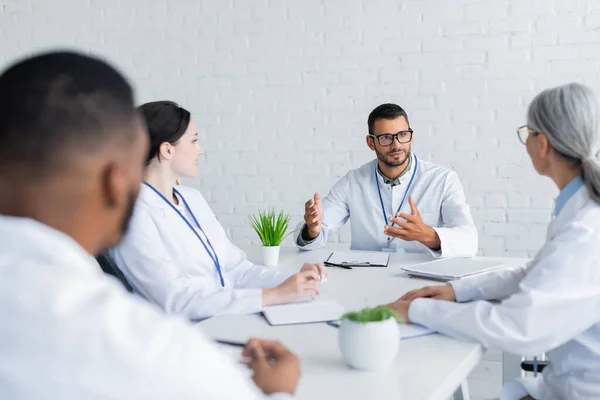 Arzt mit Brille gestikuliert im Gespräch mit verschwommenen multiethnischen Kollegen — Stockfoto