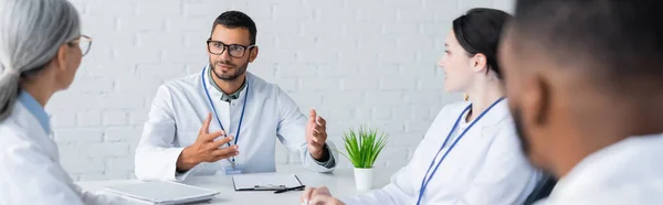 Joven médico gesticulando mientras habla con colegas multiculturales, bandera - foto de stock