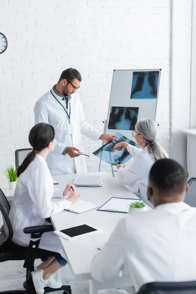 Médecin mature pointant vers les poumons radiographie pendant le conseil avec des collègues multiethniques — Photo de stock