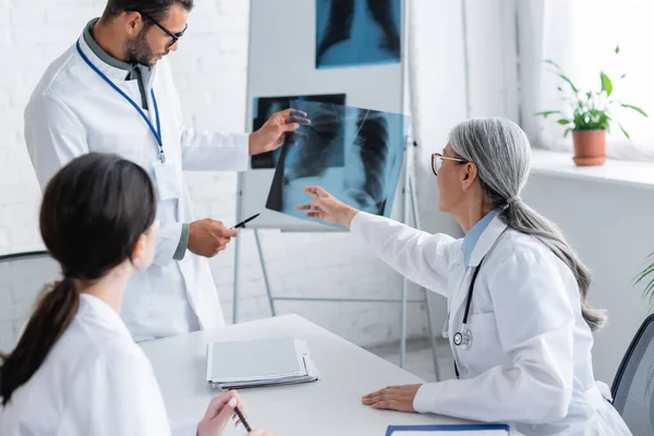 Mature asian doctor pointing at lungs x-ray during meeting with young colleagues — Stock Photo