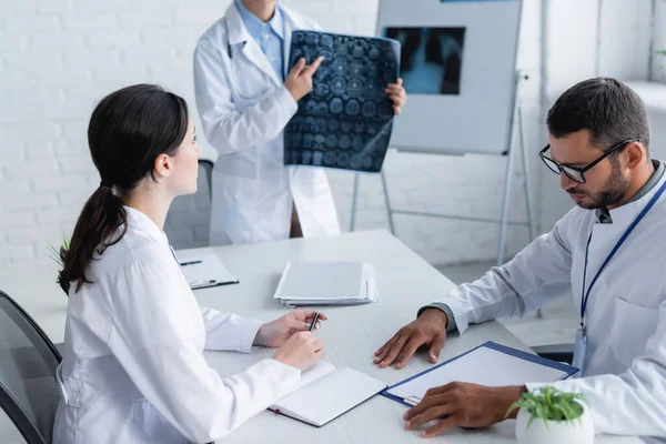 Doctor apuntando a la resonancia cerebral cerca de colegas que trabajan con documentos - foto de stock