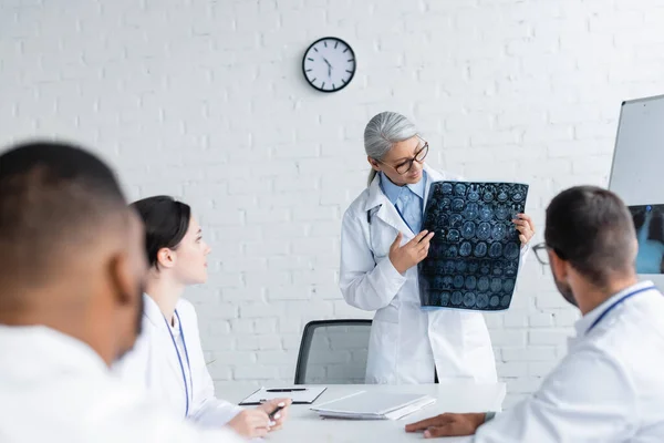 Mature asian physician showing head mri scan to blurred multicultural doctors — Stock Photo