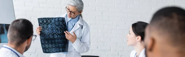 Middle aged asian doctor showing brain mri scan to blurred multiethnic colleagues, banner — Stock Photo