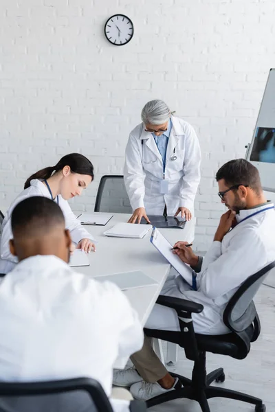 Asiatischer Arzt mittleren Alters schaut sich Röntgenbilder in der Nähe multiethnischer Kollegen an, die während des Rates schreiben — Stockfoto