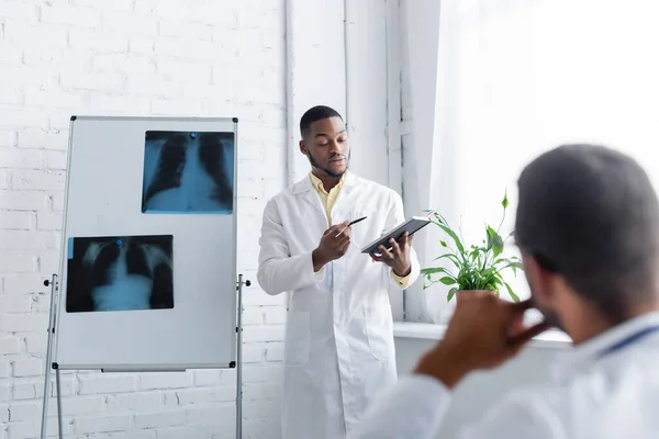 Afrikanisch-amerikanischer Arzt zeigt auf Notizbuch in der Nähe von Lungen-Röntgenaufnahmen und verschwommener Kollege — Stockfoto