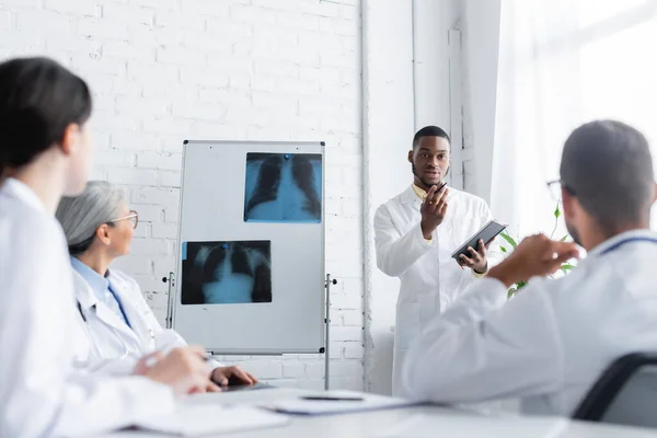 Médico afroamericano gesticulando mientras habla con colegas borrosos cerca del rotafolio con radiografías pulmonares - foto de stock