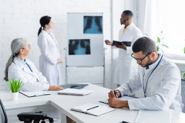 Joven médico escribiendo en el portapapeles mientras borrosa colegas multiétnicos hablando cerca de los pulmones rayos X - foto de stock