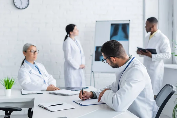 Junger Arzt schreibt in Notizbuch, während multikulturelle Kollegen nahe der Lunge Röntgenaufnahmen auf verschwommenem Hintergrund machen — Stockfoto