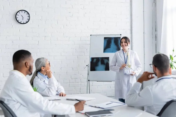 Médico sonriente de pie cerca del rotafolio con radiografías pulmonares y mostrando el modelo de columna vertebral a colegas multiétnicos - foto de stock