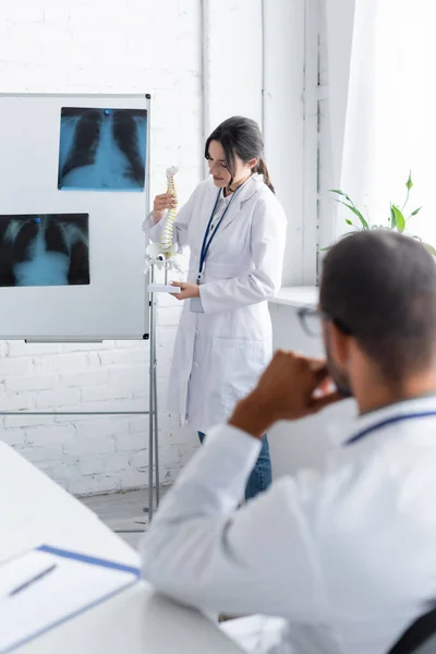 Jovem médico segurando espinha modelo puro flip chart com raios-x e colega borrado — Fotografia de Stock