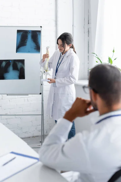 Joven médico sosteniendo modelo de columna vertebral nítido rotafolio con rayos X y colega borroso - foto de stock