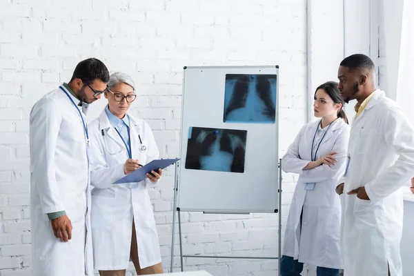 Médico asiático de mediana edad apuntando al portapapeles cerca de los pulmones fluorografía y colegas multiétnicos - foto de stock