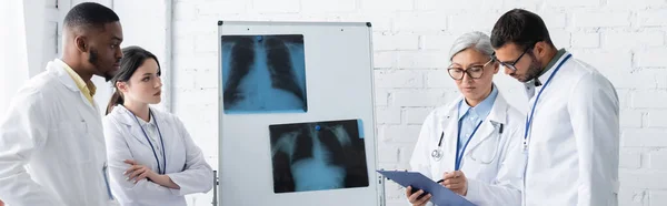 Maduro asiático médico apuntando en portapapeles cerca de flip chart con pulmones rayos X y jóvenes multiétnicos colegas, bandera - foto de stock