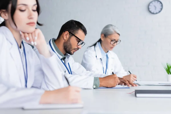 Young physician in eyeglasses writing near multiethnic colleagues in meeting room — Stock Photo