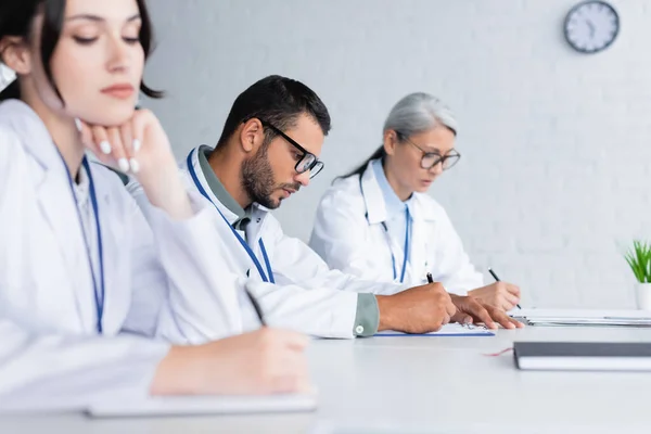 Joven médico en anteojos escribiendo cerca de colegas multiétnicos en la sala de reuniones - foto de stock