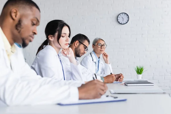 Blurred african american doctor writing near colleagues during council in hospital — Stock Photo
