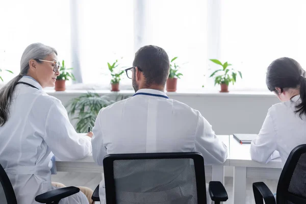 Rückansicht junger Ärzte, die in der Nähe eines reifen asiatischen Arztes im Besprechungsraum sitzen — Stockfoto