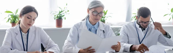 Médico reflexivo tocando la cabeza con la pluma mientras trabaja cerca de colegas multiétnicos, pancarta - foto de stock