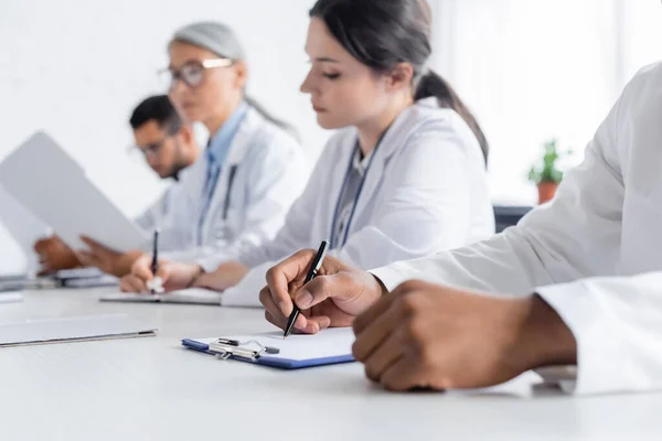 Afro-américain médecin écrit sur presse-papiers près flous collègues multiethniques en salle de conférence — Photo de stock