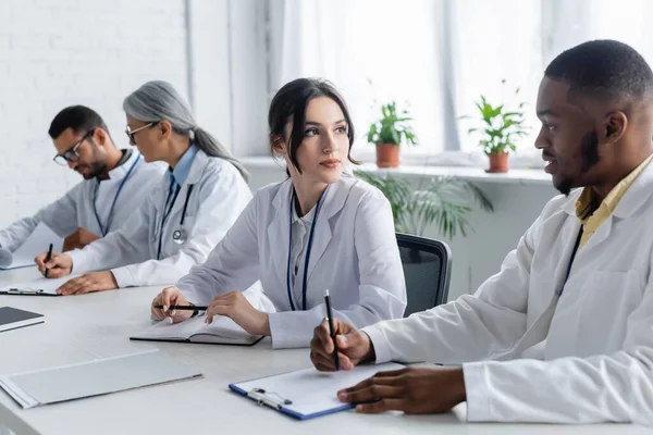 Interracial doctors looking at each other near blurred colleagues — Stock Photo