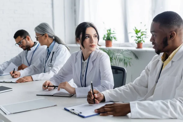 Médicos interraciales mirándose unos a otros cerca de colegas borrosos - foto de stock