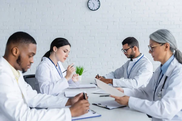 Médicos multiculturales sentados en el escritorio durante el consejo médico en la clínica - foto de stock